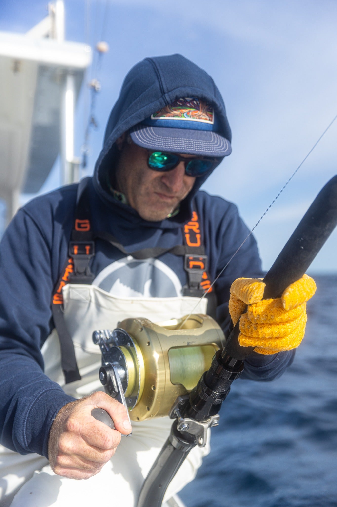 A fisherman aboard a boat working a fishing rod and reel, wearing yellow gloves